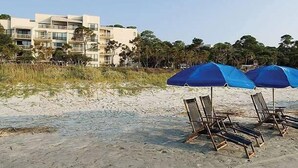 A beach with umbrellas and lounge chairs, a building with balconies, and trees in the background. - Hilton Head | Family Island Stay | Pool + Beach | 2BR Condo (Hilton Head Island)