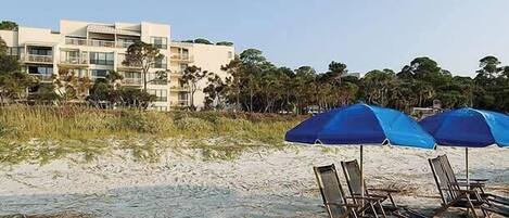 A beach with umbrellas and lounge chairs, a building with balconies, and trees in the background.
