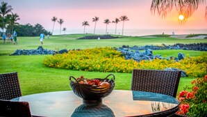 Une table à manger avec un panier de fruits au premier plan, une vue sur un terrain de golf et un coucher de soleil en arrière-plan.