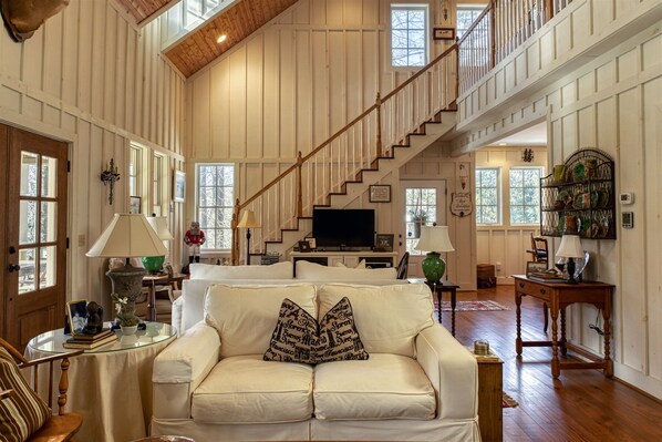 Ein geräumiges Wohnzimmer mit hoher Decke, Holzbalken, einer Treppe, einem Fernseher und einer gemütlichen Sitzecke mit einem weißen Sofa und dekorativen Kissen.