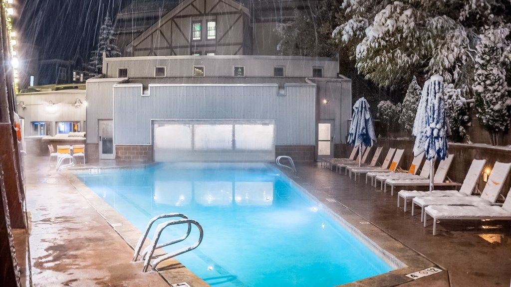 Una piscina con sillas de descanso y sombrillas, un área techada con garaje y un edificio con tejado a dos aguas.