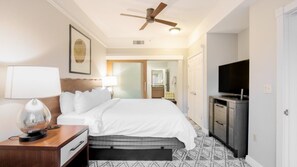 Quarto de hotel com cama, mesa de cabeceira, abajur, ventoinha no teto, televisão e banheiro visível por uma porta aberta.