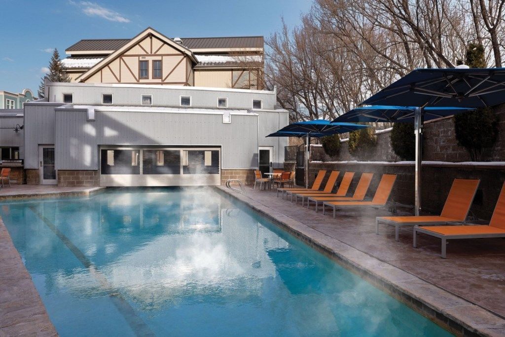 Une piscine avec des chaises longues et des parasols, un bâtiment au design moderne et un ciel dégagé.