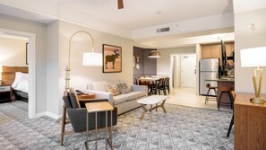Un salon moderne avec un canapé, une table basse et une lampe. On y trouve également un coin repas avec des chaises et une table. Une cuisine équipée d’un réfrigérateur et d’une cuisinière est visible au fond.