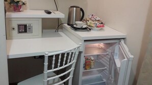 Une petite cuisine équipée avec un plan de travail blanc, un petit réfrigérateur, une chaise blanche et un téléviseur fixé au mur.