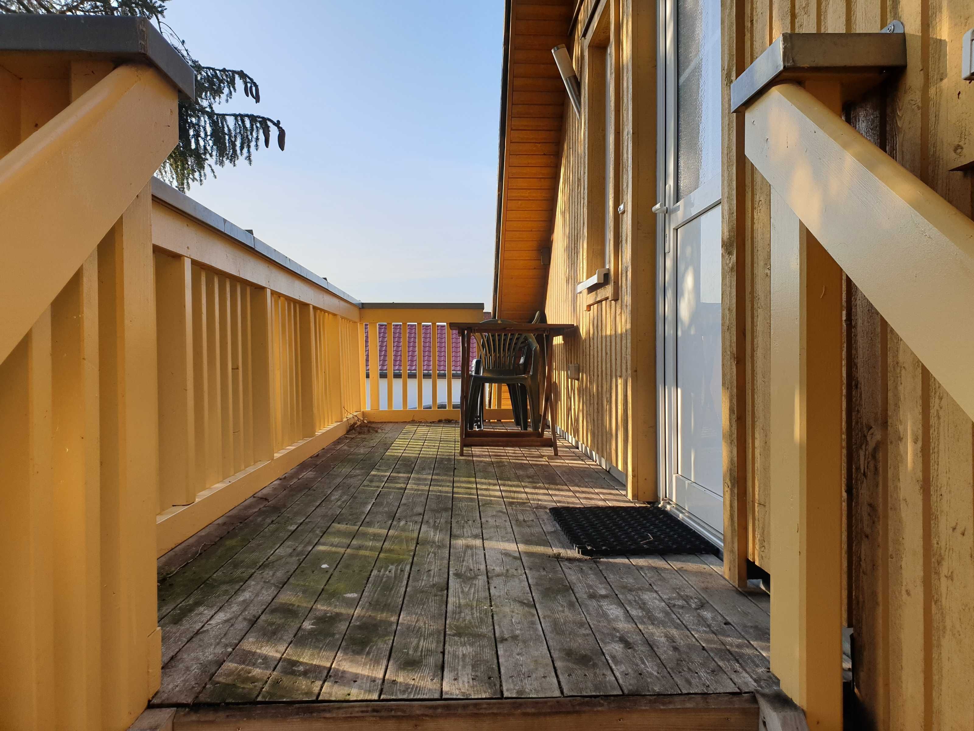 Ett trädäck med ett räcke, en överbyggd sittplats och en dörr som leder in i en byggnad.