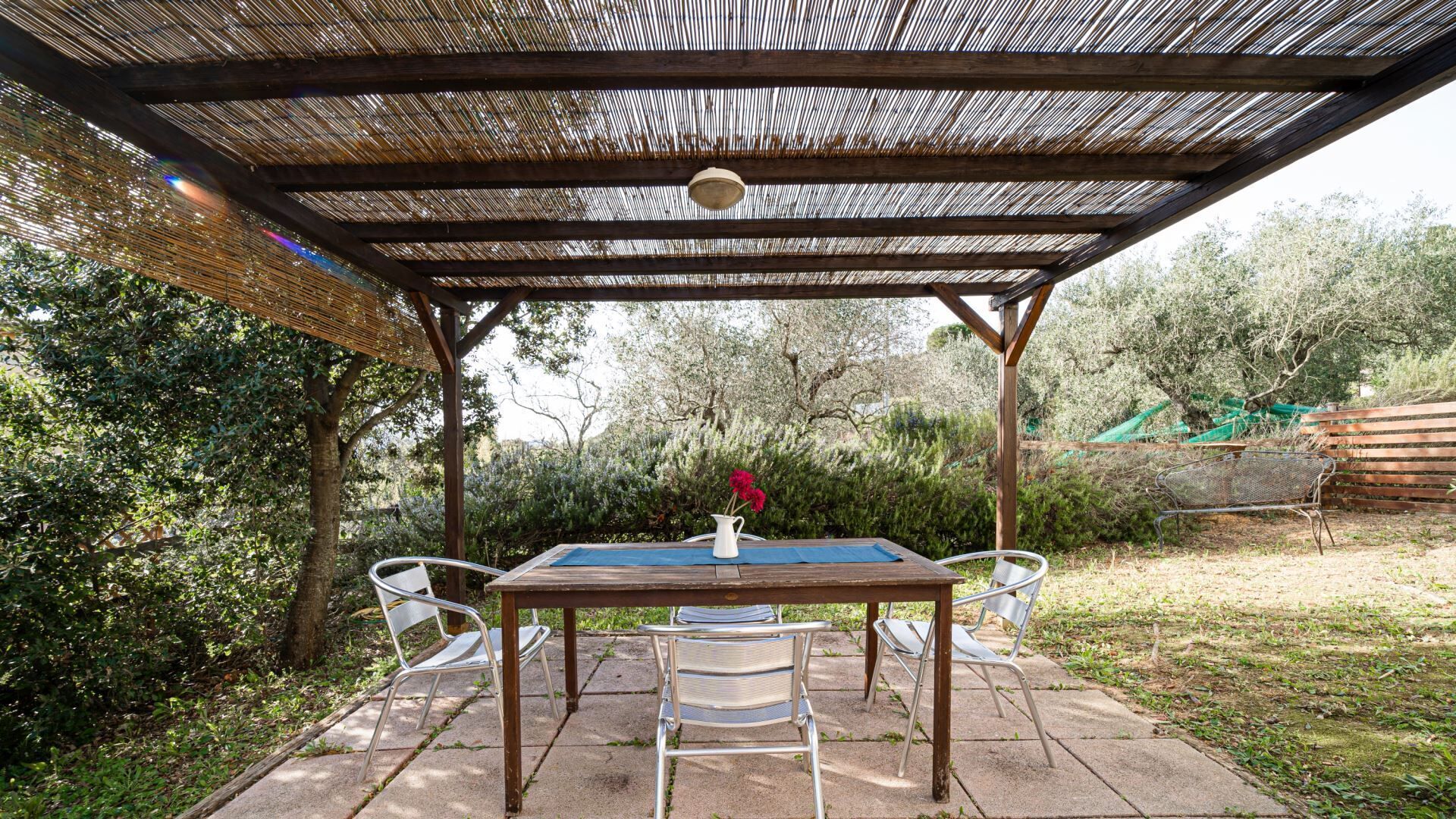 An outdoor patio with a wooden table and four chairs, a white pitcher with flowers, and a pergola overhead.