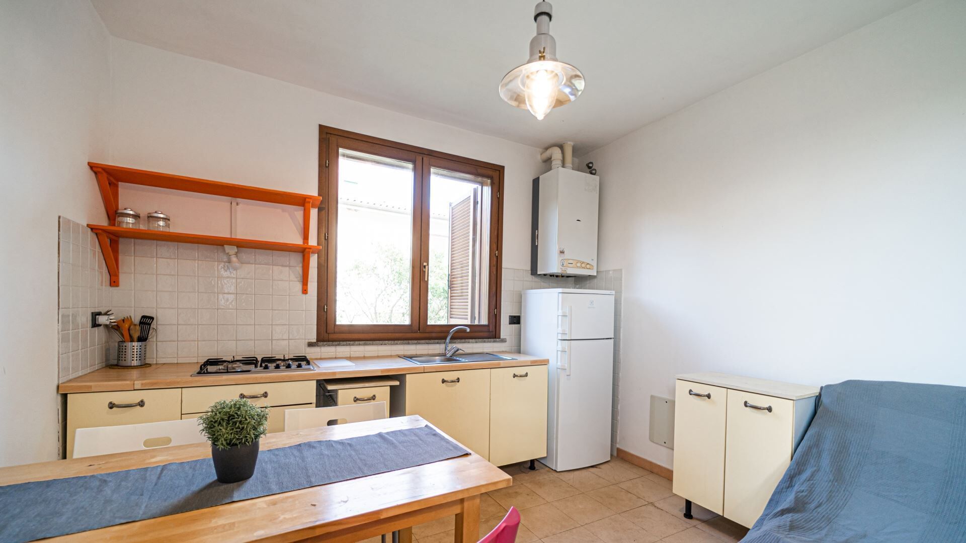 Eine kleine Küche mit Gasherd, Spüle und weißen Schränken. Ein Fenster mit Jalousien, eine Deckenleuchte und eine kleine Pflanze auf dem Tisch sind vorhanden.