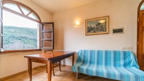 Una habitación con una mesa de madera, un sofá azul a rayas, un ventanal con vistas a la vegetación, una lámpara fijada a la pared y un cuadro enmarco.