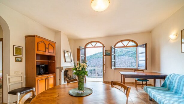 Un área de comedor con una mesa de madera, sillas y un sofá. Hay una ventana con un persiano abierto, un jarrón con flores y un mueble con estantes.