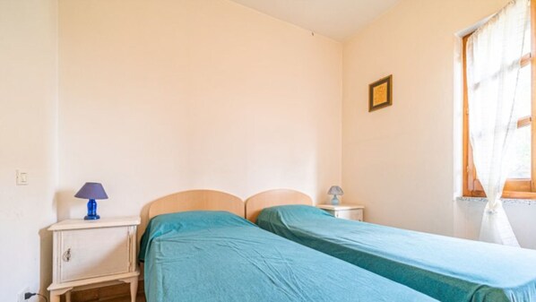 A bedroom with two single beds, a nightstand with a lamp, a small table, and a window with curtains.
