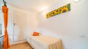 Une petite chambre avec un lit simple, une porte blanche, un rideau orange, une couvre-lit beige et un tableau représentant un tournesol accroché au mur.