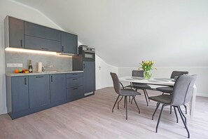 Une cuisine moderne avec des meubles de couleur sombre, un four encastré et un coin repas comprenant une table blanche et des chaises grises.