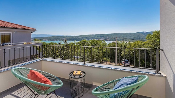 A balcony with two wicker chairs, a small table, and a view of a lake and mountains.