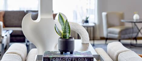 A modern living room with a white abstract sculpture, a potted plant, and a tray with books titled "Foraged Flora," "Luxury Toys," and "Classic Cars."