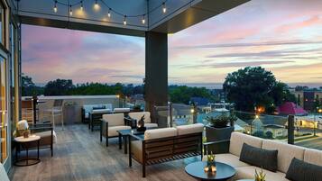 Terraço no telhado com mesas ao ar livre, luzes de festa e vista para a cidade ao entardecer.