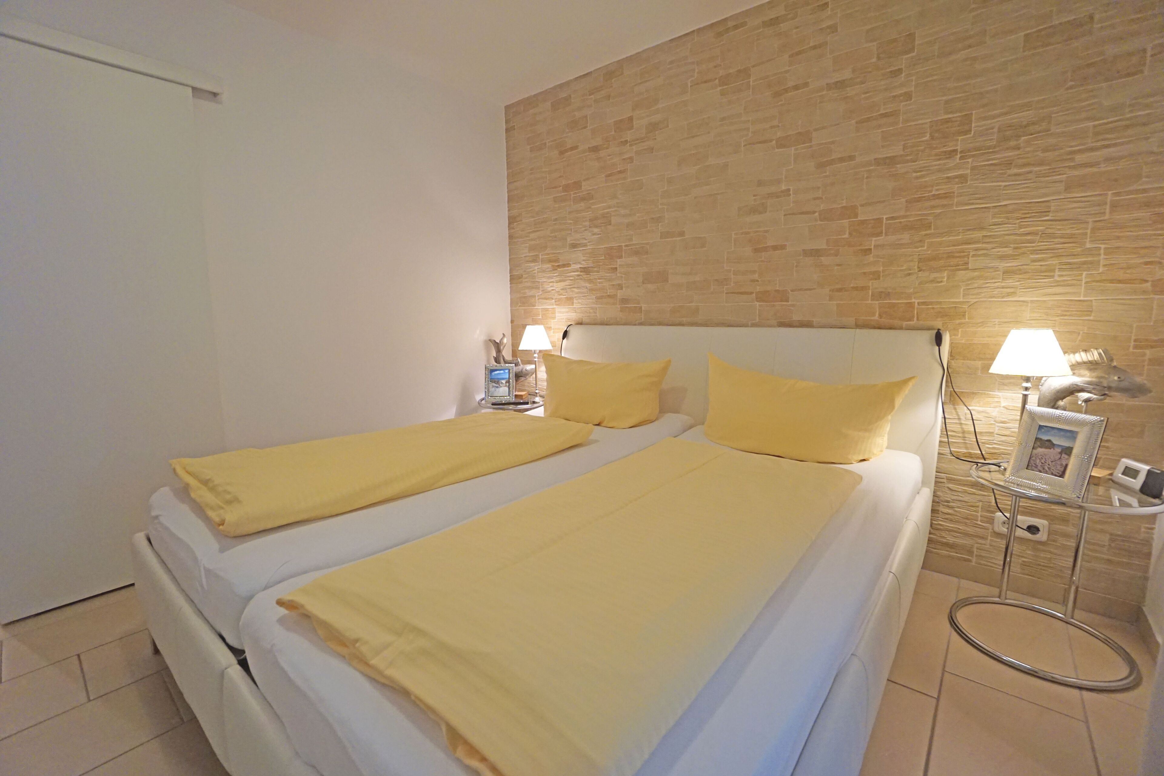 A double bed with yellow bedding, a bedside table with a lamp, and a wall with a brick pattern.