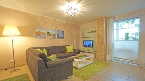 Ein Wohnzimmer mit grauem Sofa, grünen Kissen, einem weißen Couchtisch, einer Stehlampe und einem Fernseher auf einem Schrank.