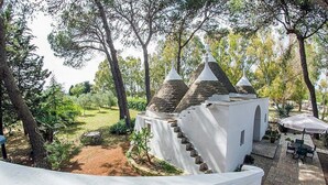 Un bâtiment blanc doté d’un escalier et d’une petite annexe, entouré d’arbres et d’un patio aménagé avec du mobilier d’extérieur.