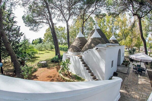 A white building with a staircase and a small annex, surrounded by trees and a patio area with outdoor furniture.