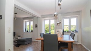 A modern dining area with a wooden table and grey upholstered chairs, large windows with a view of greenery, and pendant lighting. - Haus Düne 9, Deutschland (Hörnum (Sylt))