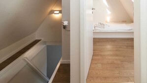 A narrow hallway leading to a bedroom with a sloped ceiling, a wooden floor, and a small bedside table with a lamp. - Haus Düne 9, Deutschland (Hörnum (Sylt))
