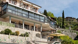Una amplia piscina con solárium de piedra, un edificio de varios pisos con balcones, y un jardín ajardinado con árboles y arbustos.