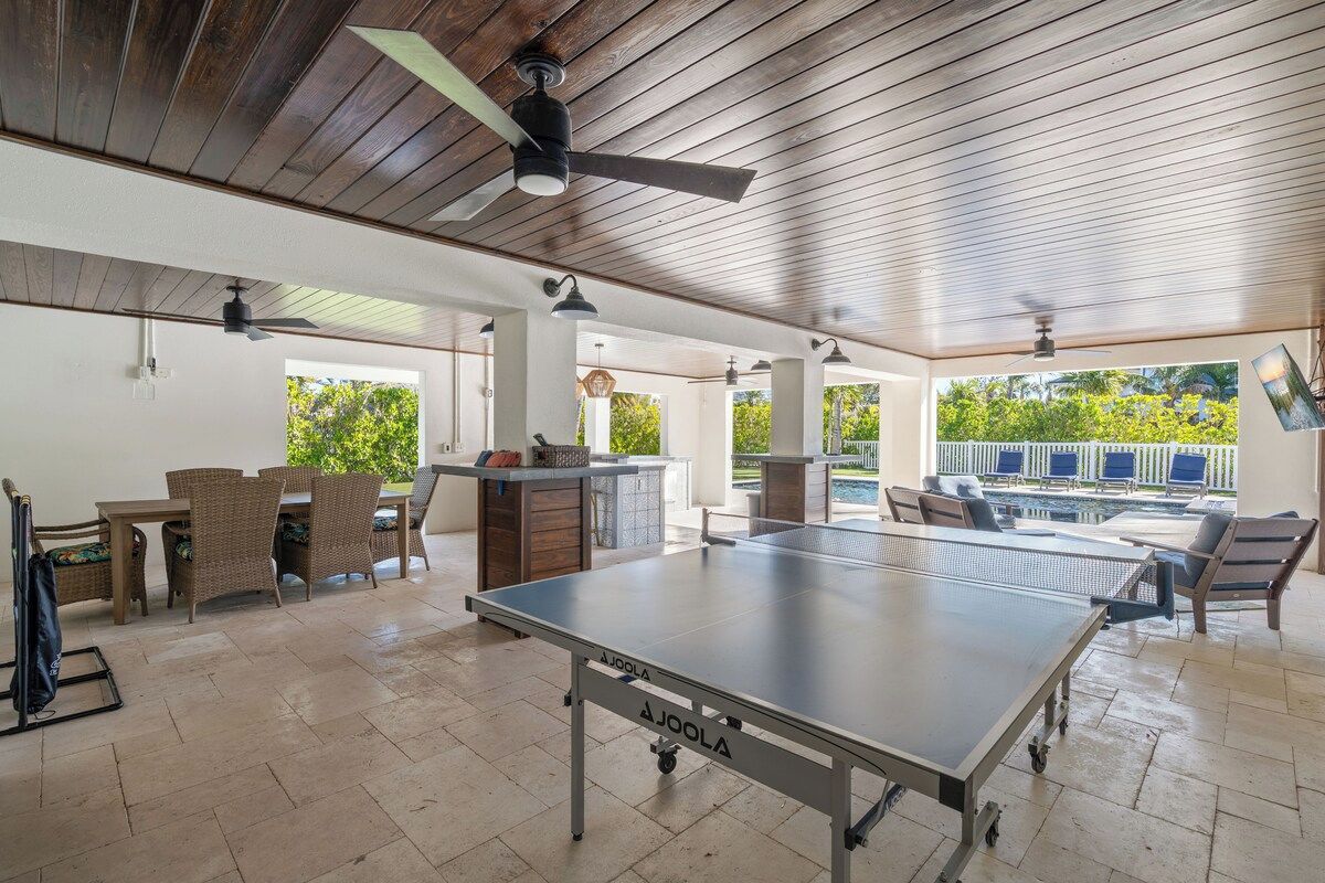 A spacious indoor area with a ping pong table, wicker furniture, ceiling fans, and a view of greenery outside.