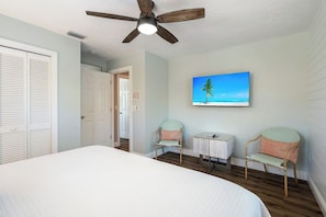 A bedroom with a bed, two chairs, a small table, a ceiling fan, and a painting of a beach scene.