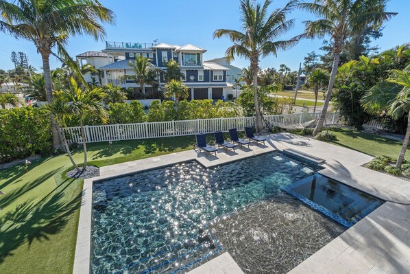A swimming pool with a clear water feature, surrounded by a white fence, palm trees, and a large house in the background.
