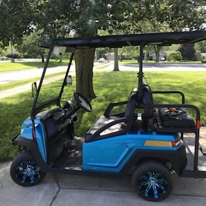 A blue golf cart parked on a sidewalk with a tree and a residential street in the background.