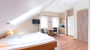 A bedroom with a wooden bed, a desk, a chair, a television, and a kitchenette in the background. - Airport Hotel One (Sylt)