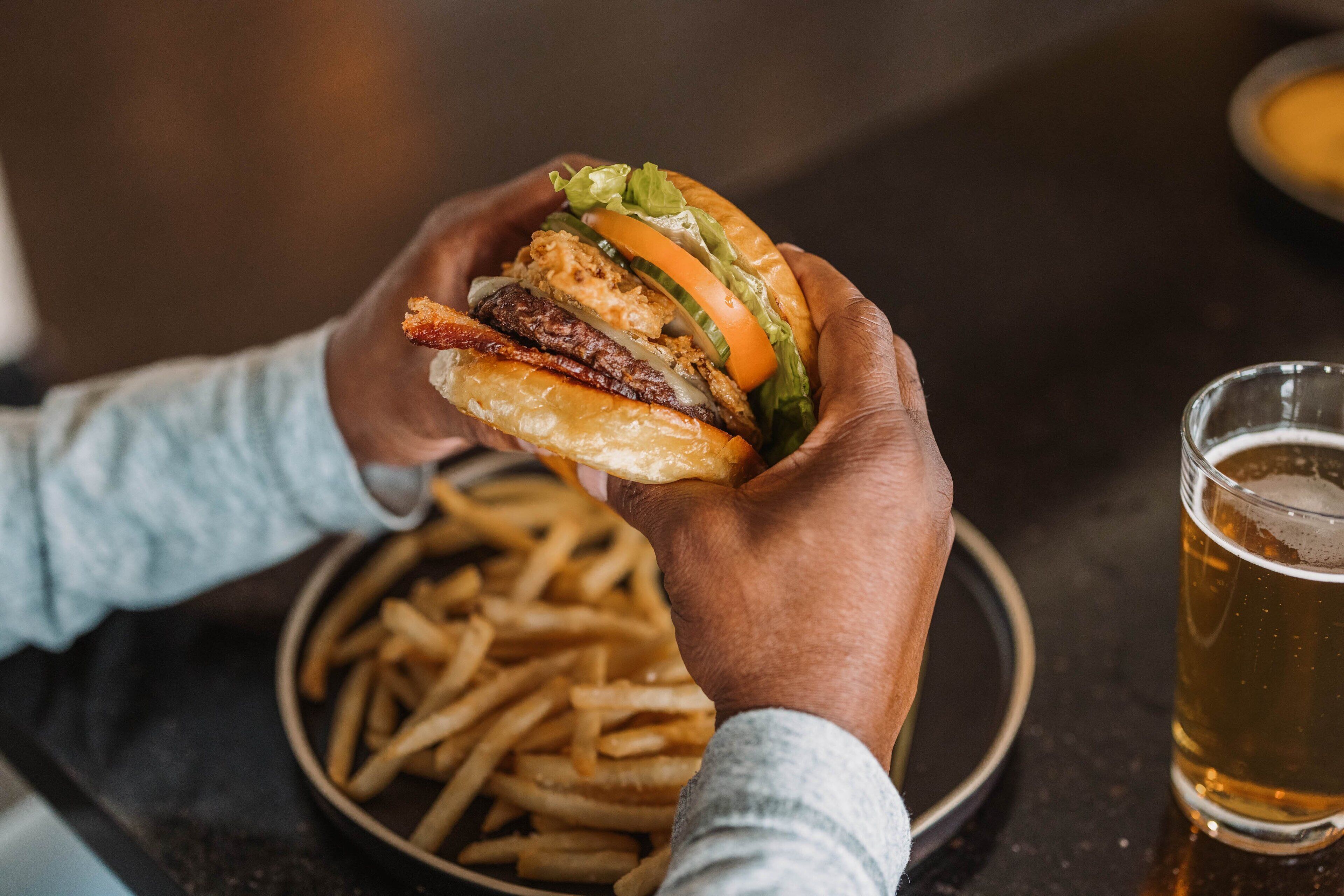 A person holding a burger with fries and a glass of beer on a table.