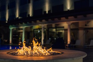 A circular outdoor fire feature with flames, surrounded by a pool and a building with columns and glass windows in the background. - Hotel Indigo Spring - Woodlands Area by IHG (Spring)