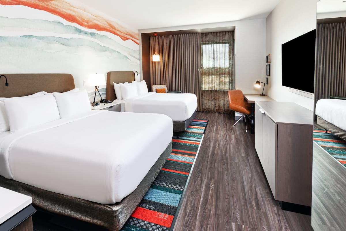 Habitación de hotel con dos camas, un escritorio, un televisor y un ventanal grande con cortinas.