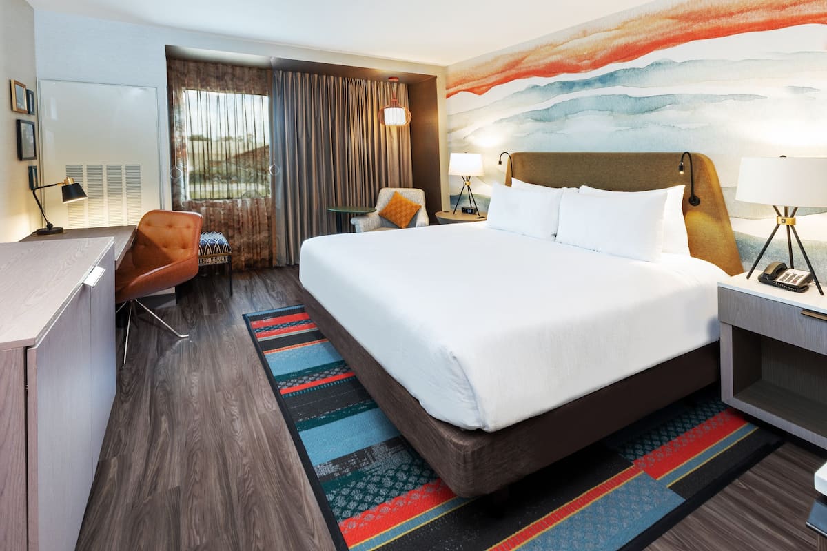 Una habitación de hotel con una cama grande, un escritorio, una silla, una mesita y un ventanal con cortinas.