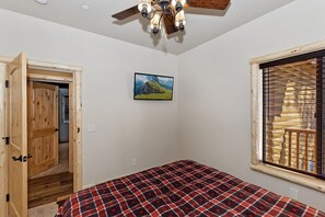 Ein Schlafzimmer mit einem Bett, einer Holztür, einem Deckenventilator, einem Fenster mit Jalousien und einem an der Wand befestigten Fernseher.