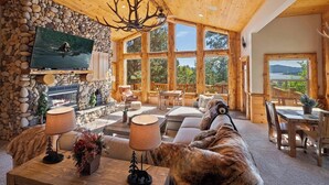A cozy log cabin interior with a stone fireplace, a television mounted on the wall, and a large window offering a scenic view.