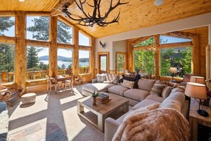 Amplia sala de estar con un sofá grande, una mesa de centro y una chimenea. Amplias ventanas ofrecen vistas al exterior.