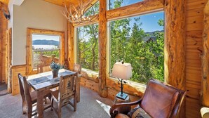 A wooden cabin interior with a dining table, chairs, a leather sofa, and a large window offering a view of greenery and mountains.