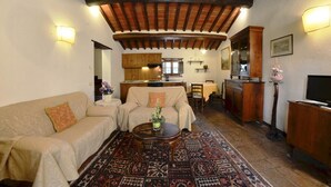 Una sala de estar acogedora con un sofá, sillones, una mesa de centro y una alfombra. Se observa una cocina al fondo con armarios de madera y un comedor.
