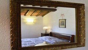 Un espejo con un marco decorativo refleja un dormitorio con cama, mesita de noche, lámpara y un cuadro en la pared.