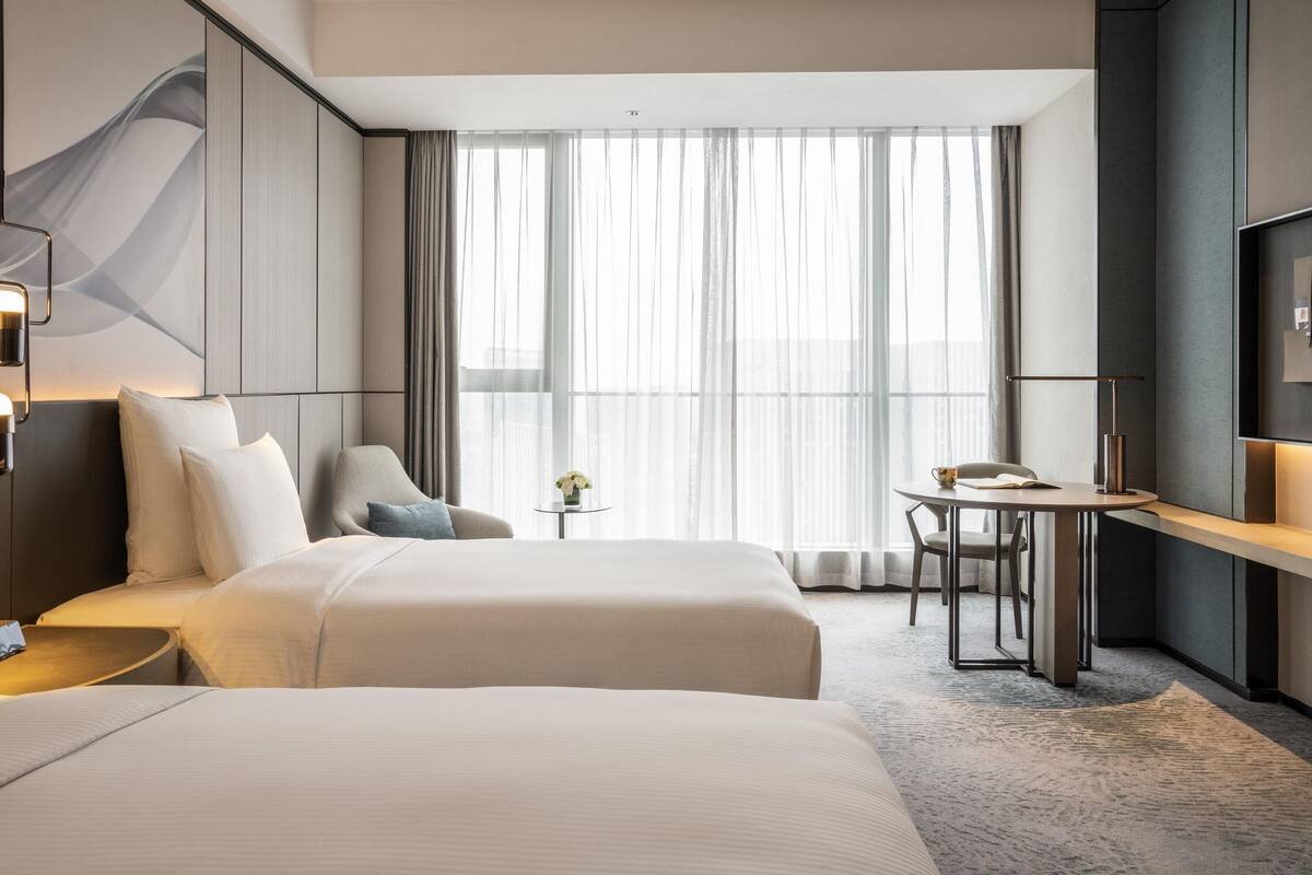 Une chambre d’hôtel avec deux lits, un bureau, une chaise, une petite table et une grande fenêtre avec des rideaux légers.