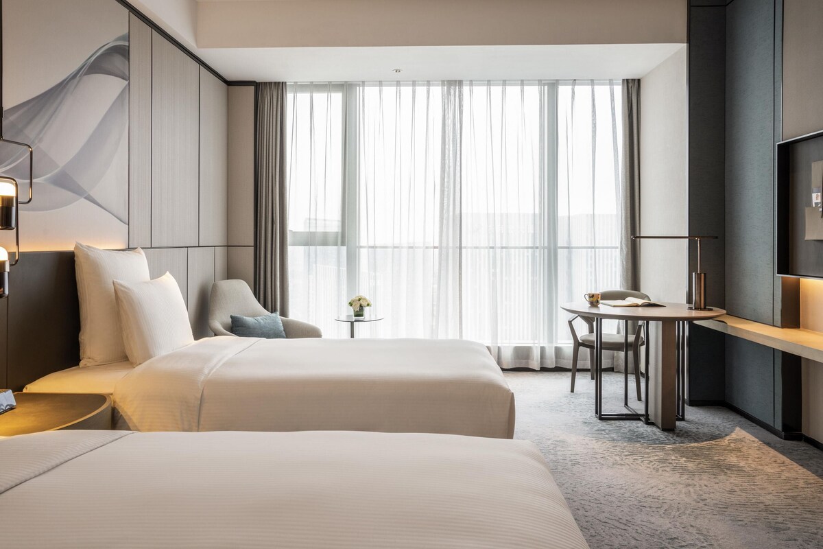 Habitación de hotel con dos camas, un escritorio, una silla, una mesita y un ventanal con cortinas de gasa.