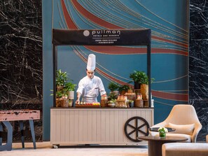 A chef in uniform at a Pullman hotel counter, with a backdrop of a wall with a string of lights and a marble surface. - Pullman Jiaxing Pinghu Excellence (Jiaxing)