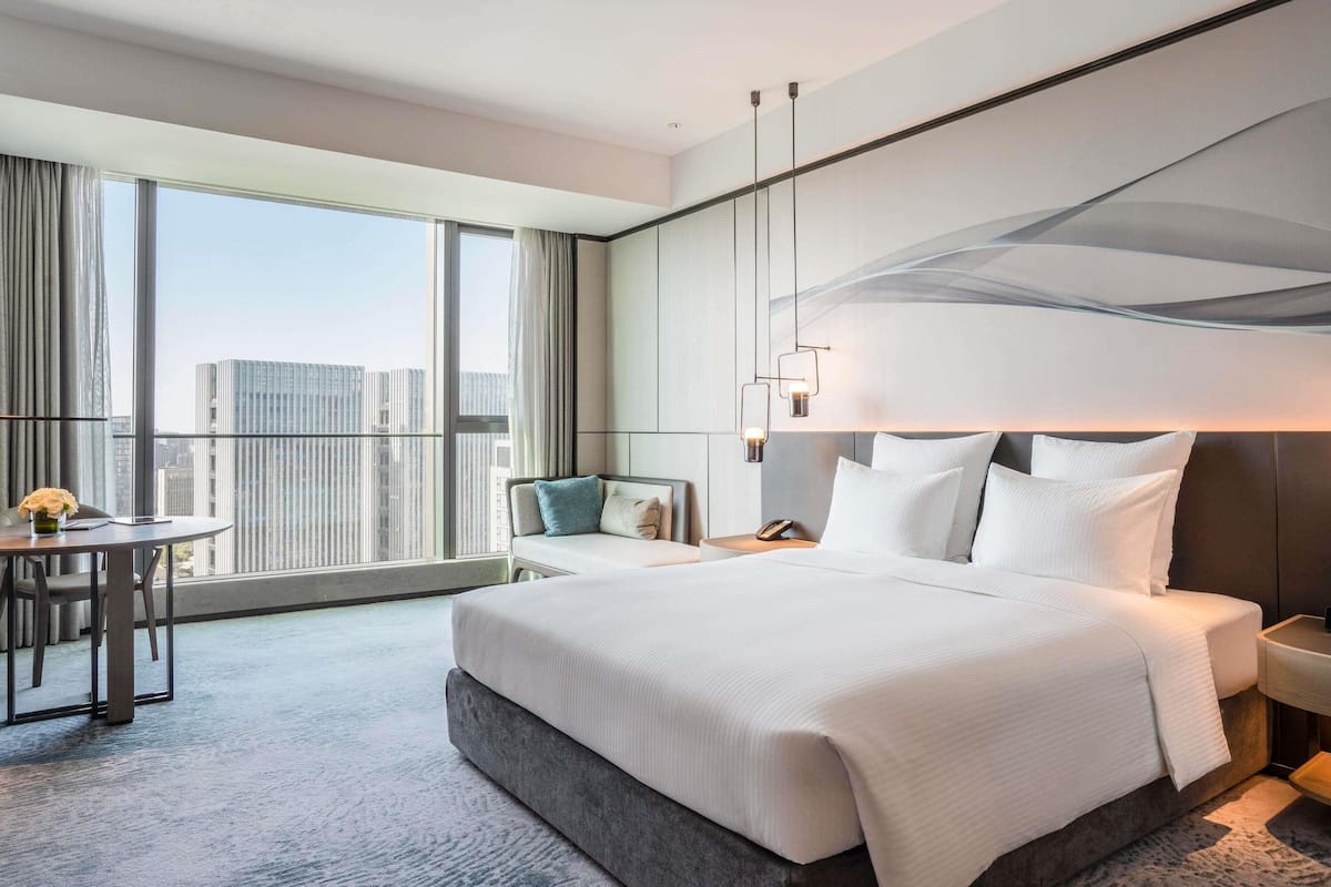 Ein modernes Hotelzimmer mit einem großen Bett, einer Sitzecke mit einem Sessel, einem kleinen Tisch und Blick auf die Stadt durch große Fenster.