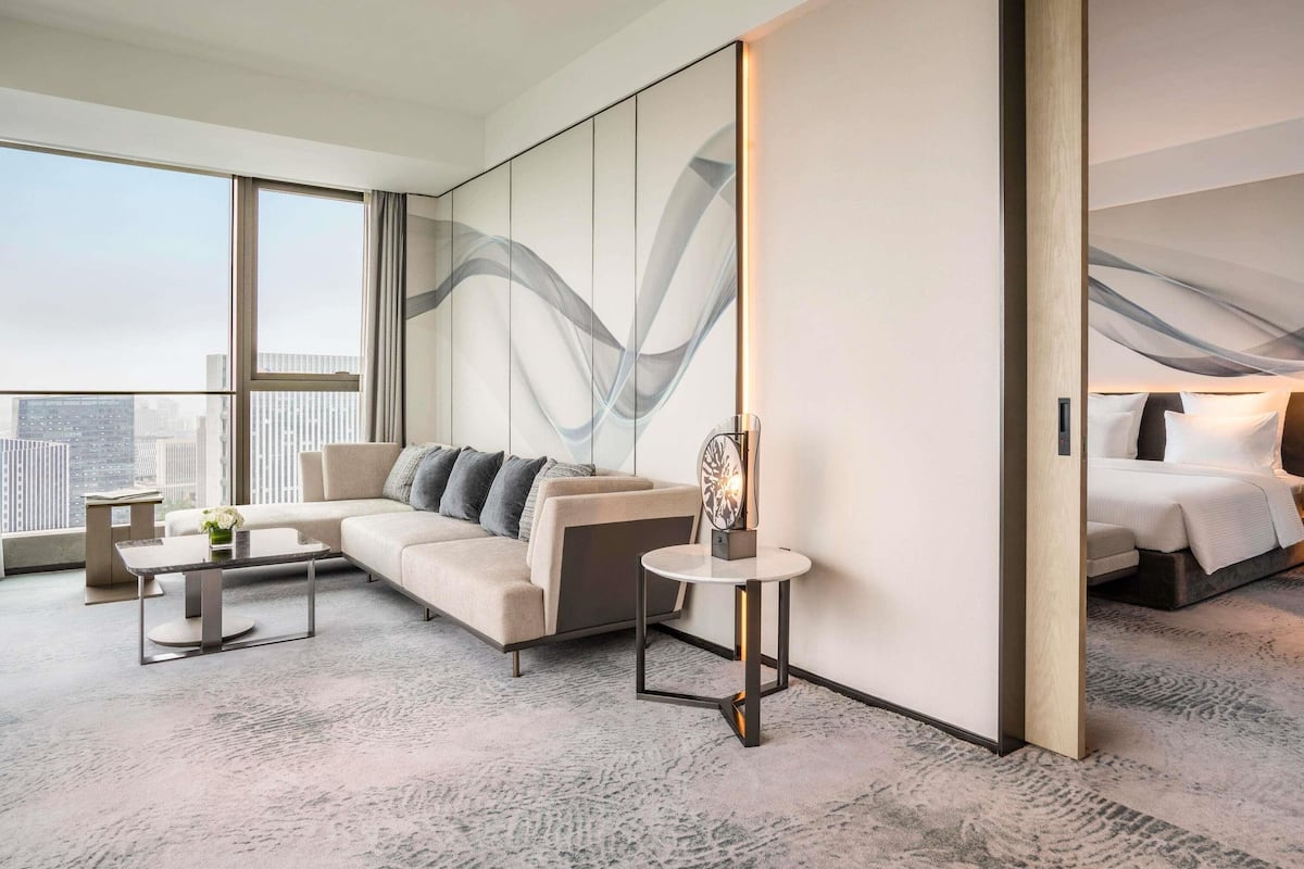A modern hotel room with a large bed, a sofa, a small table, and a large window offering a city view.