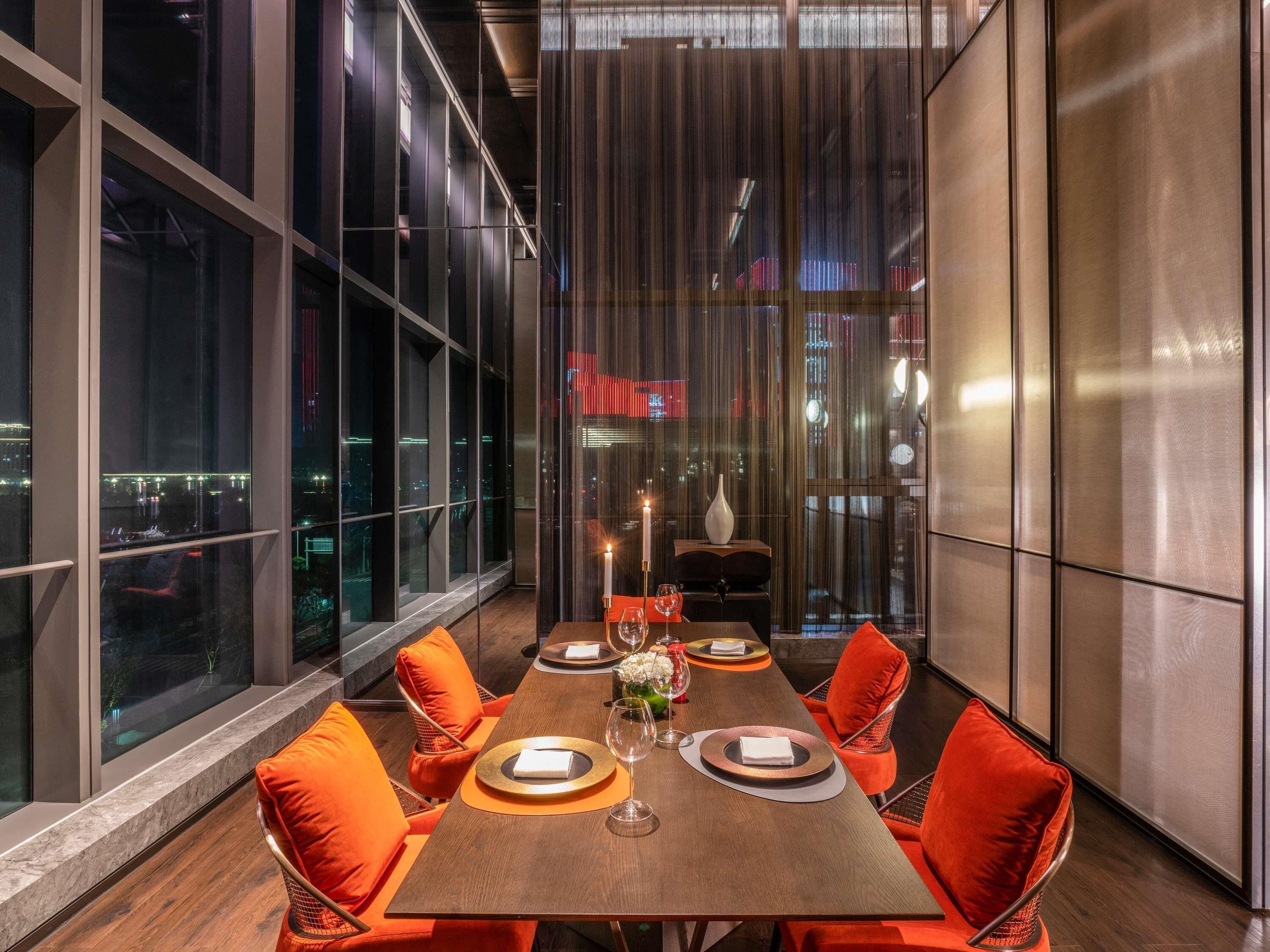 A modern dining area with a long table, orange chairs, and a city view through large windows.