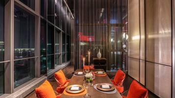 A modern dining area with a long table, orange chairs, and a city view through large windows.