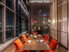 A modern dining area with a long table, orange chairs, and a city view through large windows.